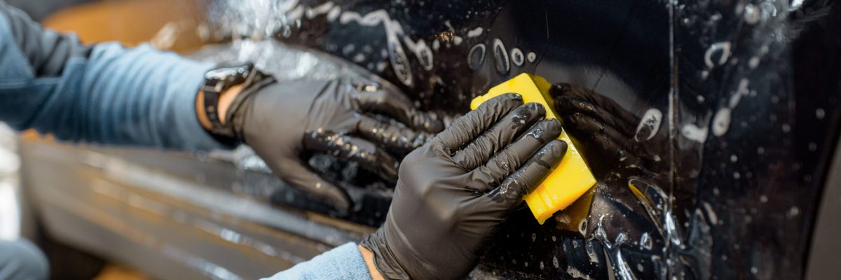 Worker sticking anti-gravel film on a car body with scrapper at the detailing vehicle workshop, close-up. Concept of car body protection with special films